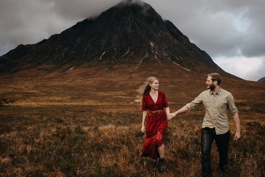 Couple walking through the grass in front of Buachaille Etive Mòr near Glencoe - wearing a boho red dress