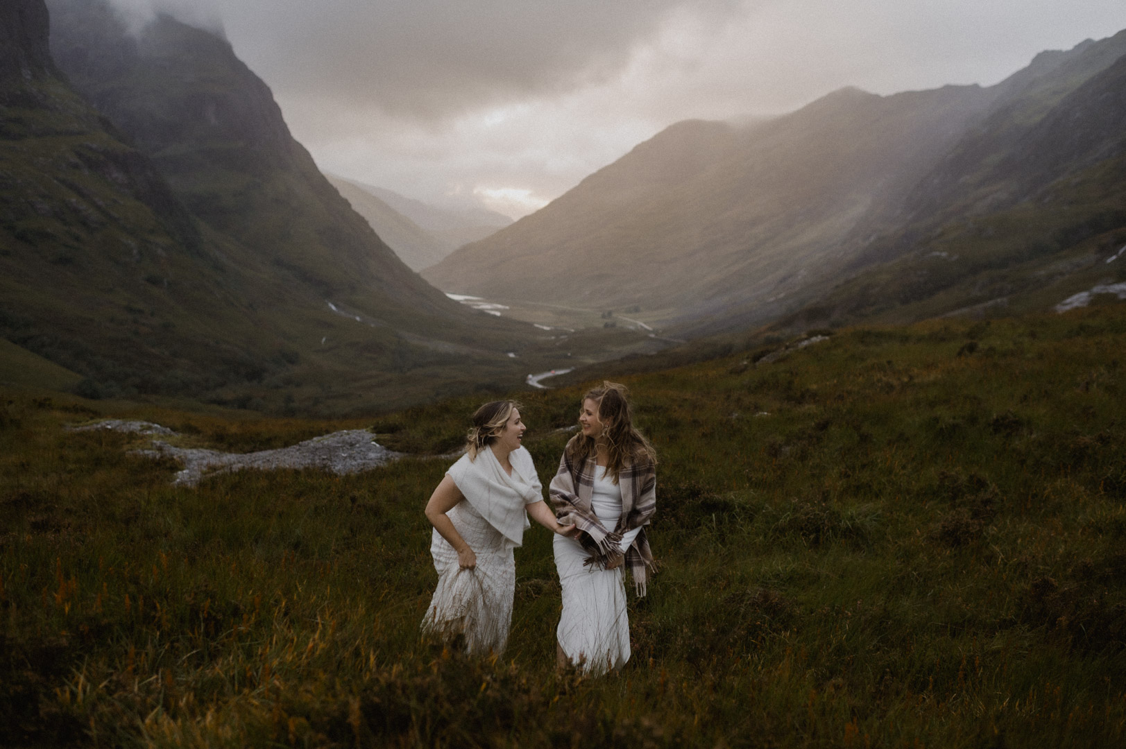 Glencoe elopement with stormy sunset - two brides laughing and climbing hill