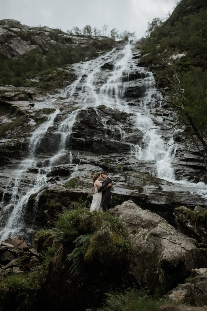 Adventurous couple cuddle at bottom of dramatic waterfall in Scottish elopement